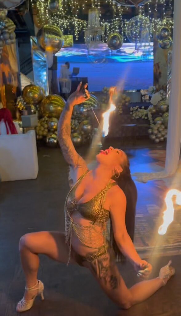A female fire performer kneels in a dramatic pose indoors, wearing a gold costume and heels, as she performs a fire-eating act under warm lights and gold balloon decorations.